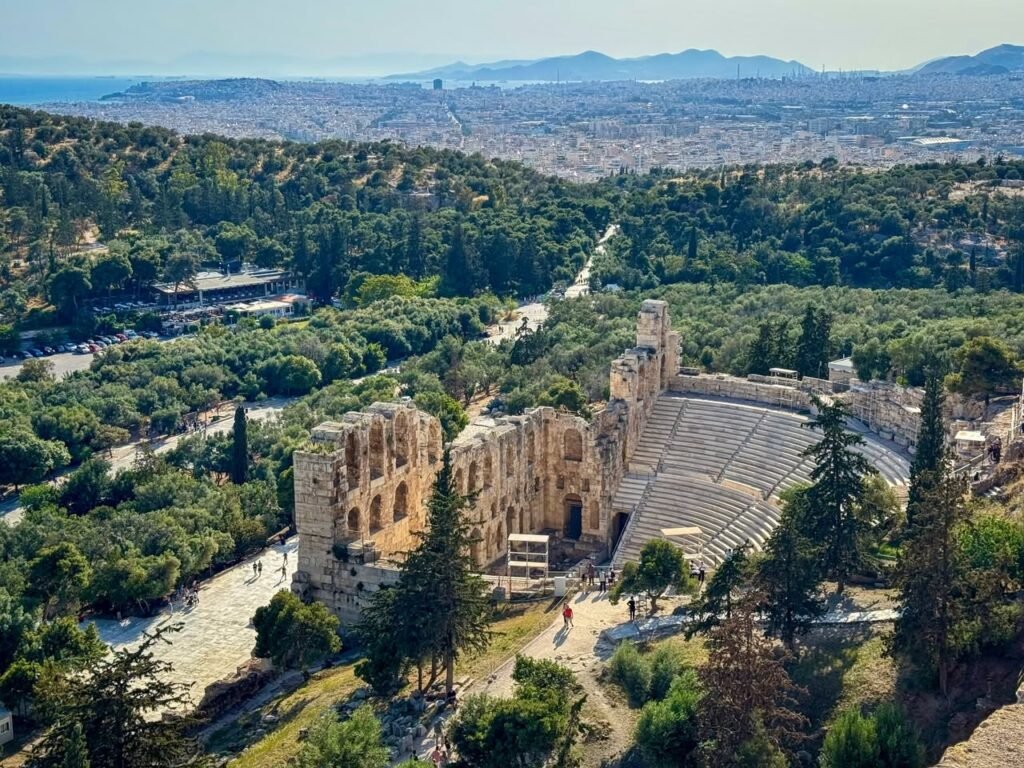 Aerial photograph of the historic Epidaurus Theatre in Greece set among dense pine trees. The view showcases the amphitheater, ancient pathways, and a panoramic vista of the surrounding landscape below.