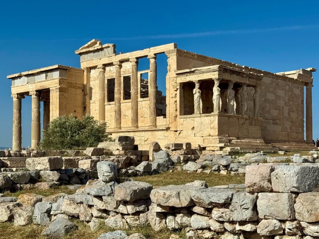 Ancient stone columns and ruins of the Acropolis of Athens under a blue sky. Parts of the Parthenon and Temple of Athena Nike are visible.