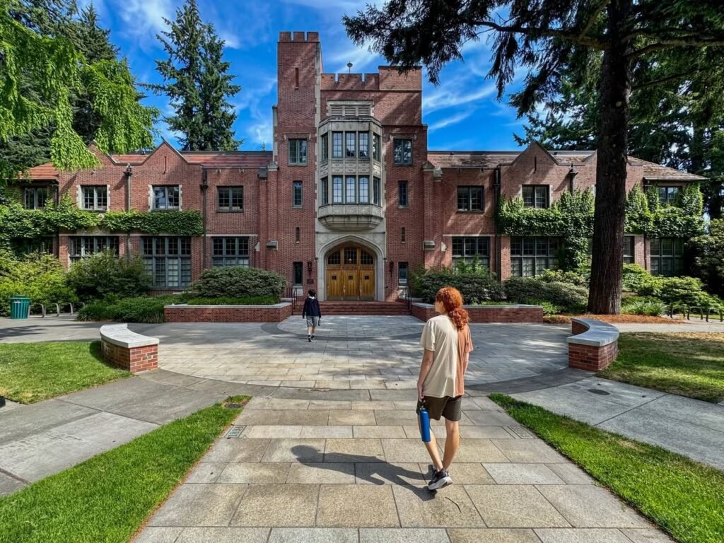 A historic red brick building with architectural details stands on a sunny day. A paved walkway and manicured gardens are featured in the Pacific Northwest.