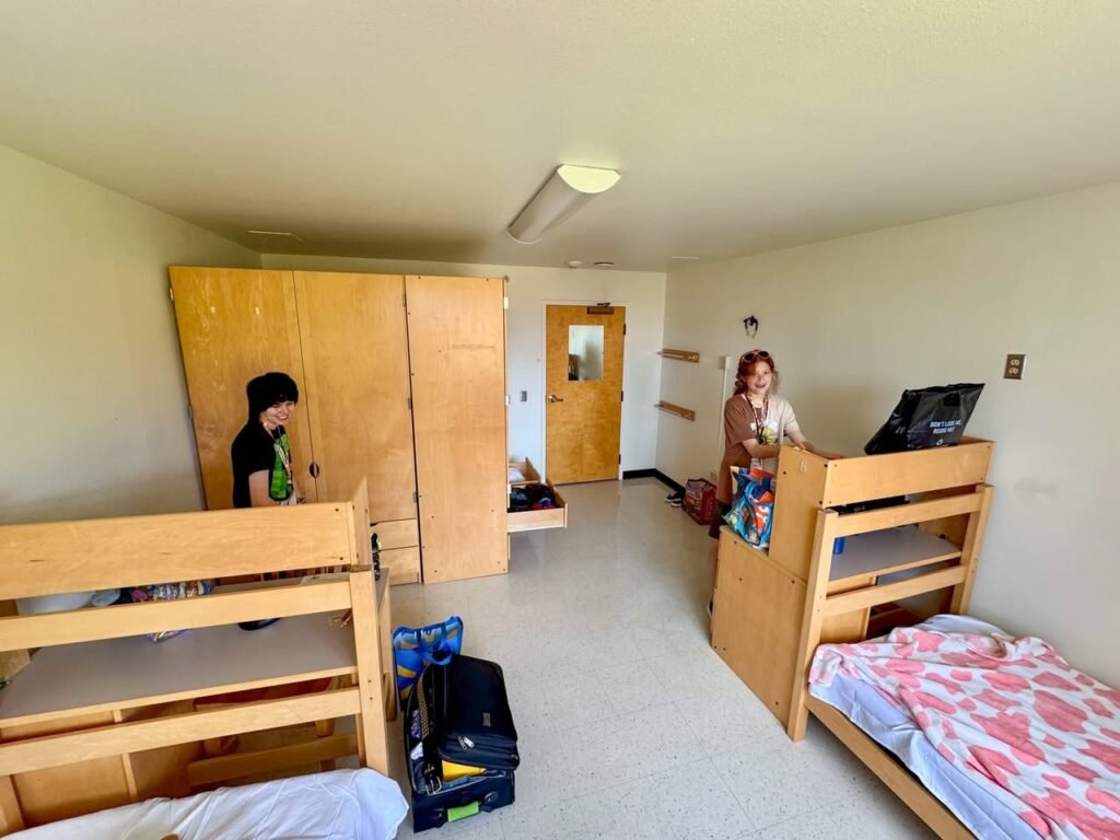 A cozy dormitory room featuring two sets of wooden bunk beds, built-in desks, and large plywood cabinets.