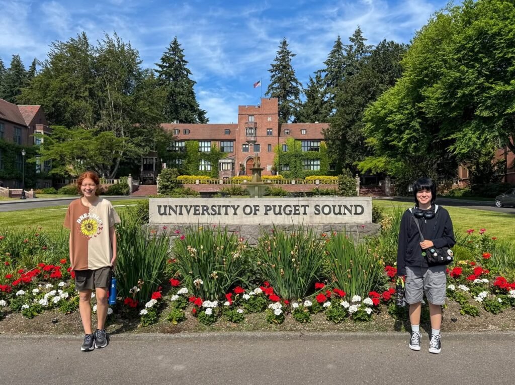 Individuals standing in front of the University of Puget Sound entrance sign, with a view of the campus architecture and background.
