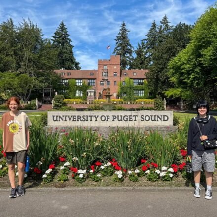 Individuals standing in front of the University of Puget Sound entrance sign, with a view of the campus architecture and background.