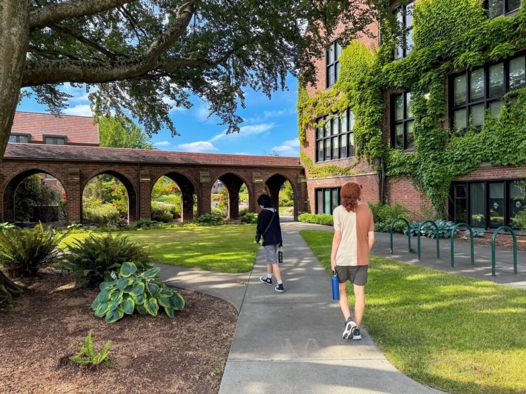 Two people walking on a concrete path across a college campus with an ivy-covered brick building on the right and open green lawns on the left.