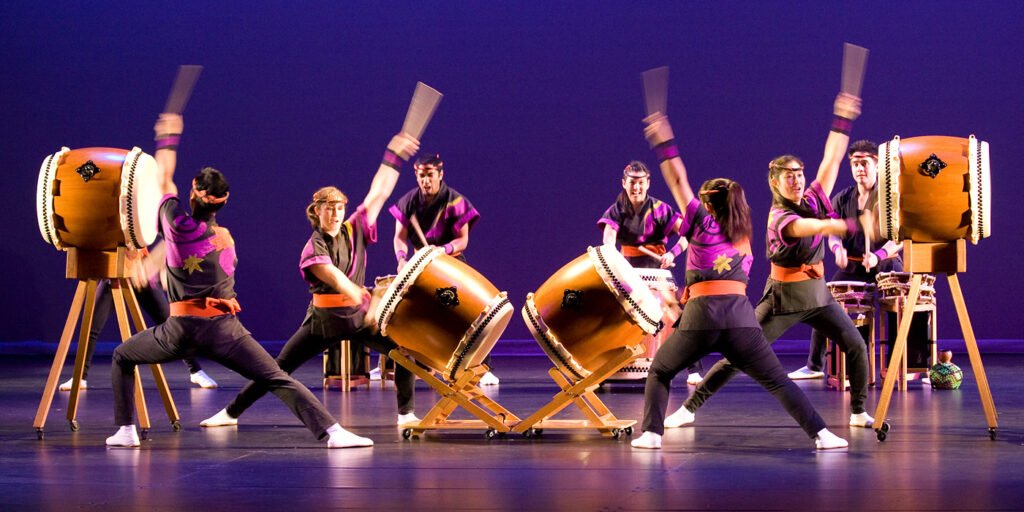 A group of performers dressed in traditional attire playing large drums on stage. They strike the drums with synchronized and dynamic movements.