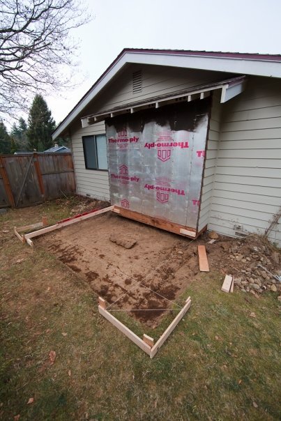 A partially constructed patio adjacent to a house with weatherproofing foil, showing wooden framing and dirt area.