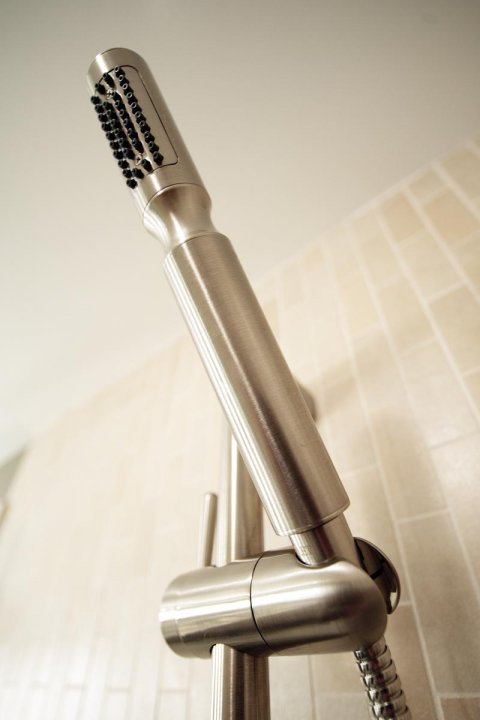 A close-up view of a modern stainless steel showerhead with a textured spray face attached to a wall-mounted fixture against a tiled background.