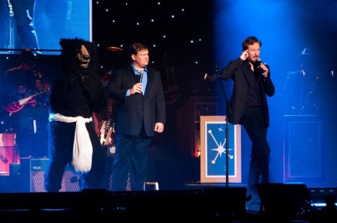 Three performers on a concert stage with a blue-lit backdrop, featuring a person in a black bear-like mask and white sash.