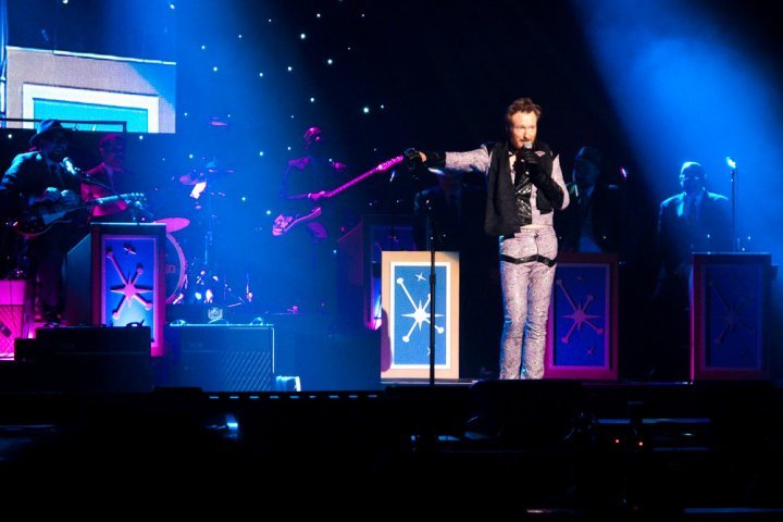 A performer on a vibrant concert stage under blue and purple lighting, with band members visible in the background.