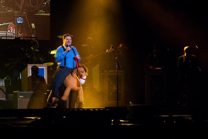 A performer in a blue shirt sings into a microphone on a concert stage illuminated by dramatic yellow lighting.