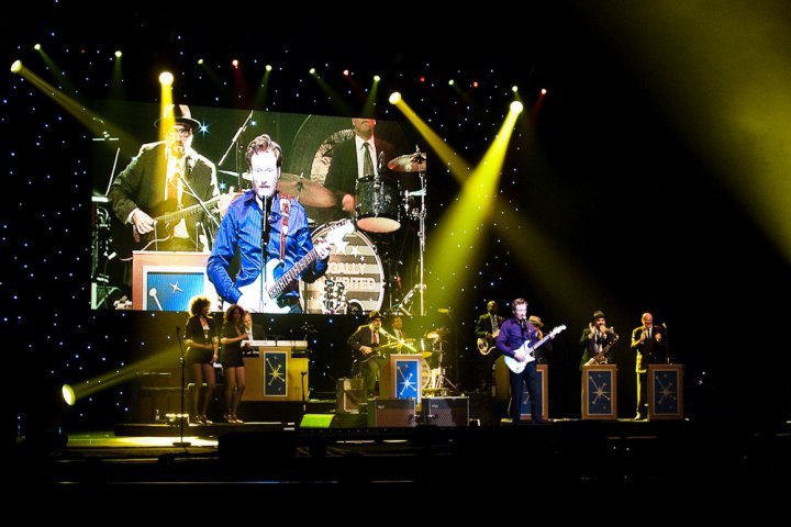 Wide view of a concert stage with musicians performing under bright yellow stage lights. A drummer is prominently silhouetted against a spotlight and a large video screen.