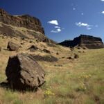 Wide view of a grassy area with scattered boulders, hills, and a clear blue sky.