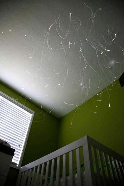 Photograph of mysterious light streaks on a bedroom ceiling above a crib near a window with blinds.