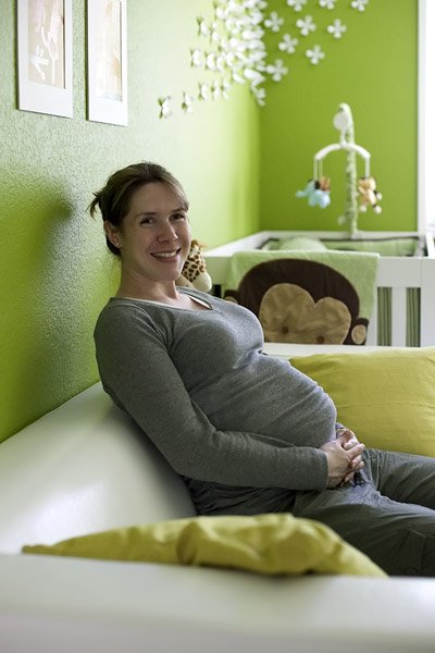 A smiling person rests on a white couch in a green nursery with a mobile and crib in the background.