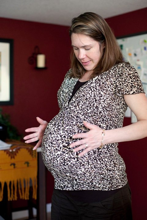 A pregnant woman cradles her belly in a cozy home setting, wearing a patterned top and black pants.