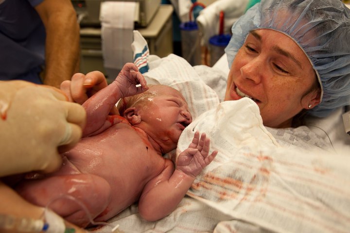 A newborn baby covered in vernix caseosa is held by a medical professional while a smiling person looks on in a hospital setting.