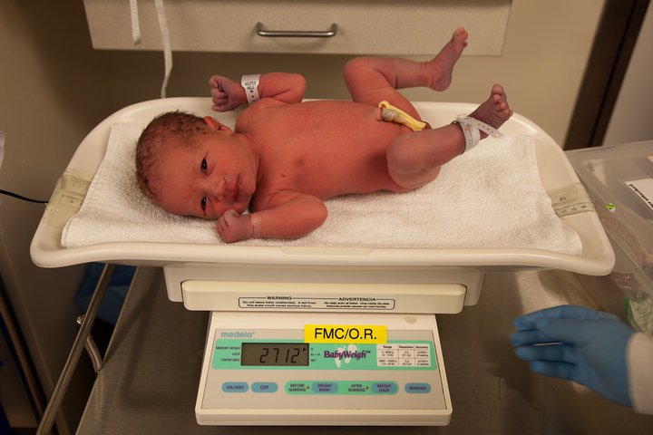 A newborn with identification bands lies on a digital medical scale in a hospital room.