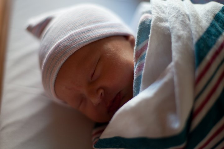 A newborn baby peacefully sleeps wrapped in a striped blanket while wearing a white hat.