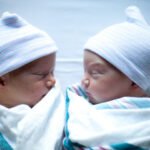 Two sleeping newborn twins wearing white striped hospital caps and wrapped in soft hospital blankets on a bed sheet.