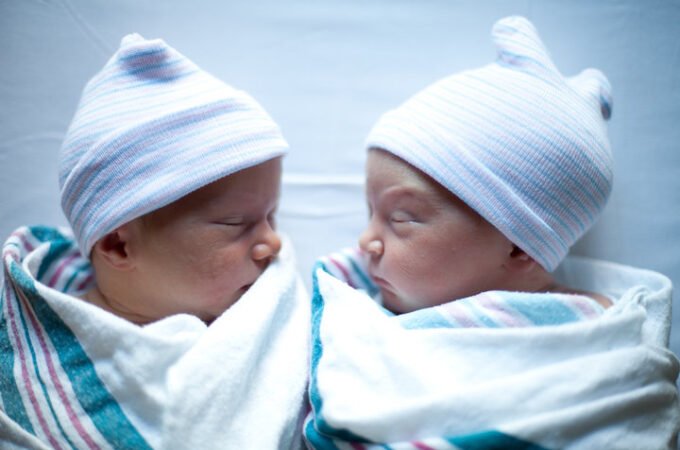 Two sleeping newborn twins wearing white striped hospital caps and wrapped in soft hospital blankets on a bed sheet.