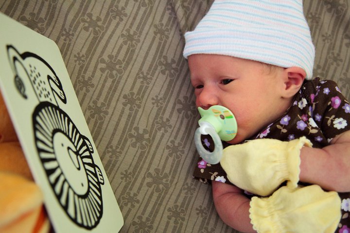 A newborn baby wearing a striped hat studies a black and white high-contrast card. The infant is holding soft yellow mittens while lying on a patterned rug.