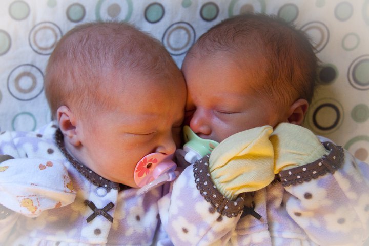 Two newborn twin babies sleeping side by side in matching outfits. Pacy pacifiers in their mouths as they rest on a patterned surface.
