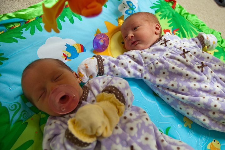 Two newborn twins lie on a colorful tropical-themed playmat with matching patterned outfits.