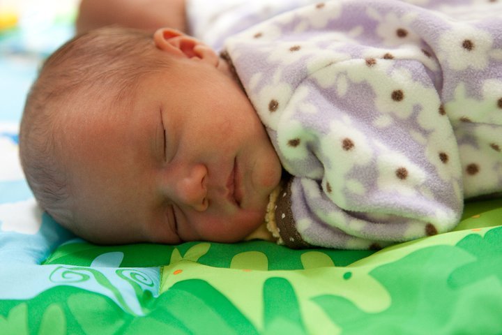 A sleeping infant wearing a patterned purple coverall rests on a green and white play mat.