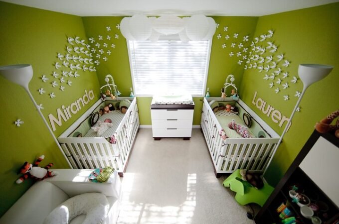 A nursery with green wallpaper featuring white stars and names. Two white cribs and a dresser are visible around a central window.