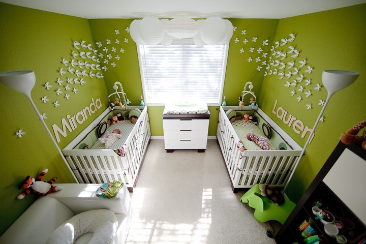 A nursery with green wallpaper featuring white stars and names. Two white cribs and a dresser are visible around a central window.