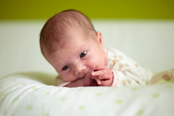 A close-up shot of a baby boy with light curly hair lying down, wearing a white polka-dotted outfit against a bright yellow wall.
