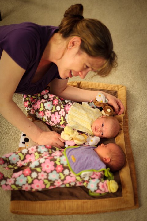 A smiling caregiver tends to two newborn babies lying on a floral blanket on the floor.