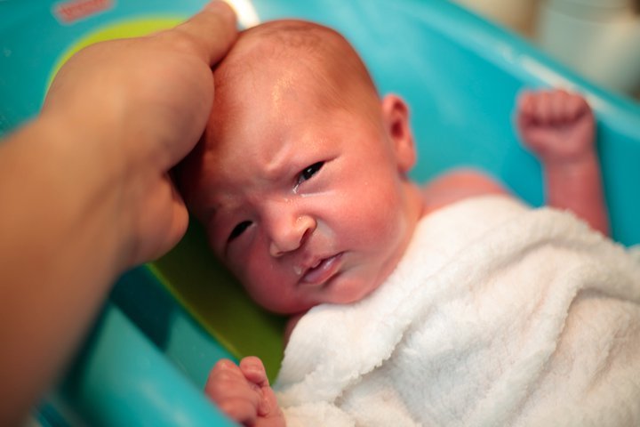 A baby, wrapped in a white towel, reaches up as an adult gently holds a comb to their head.