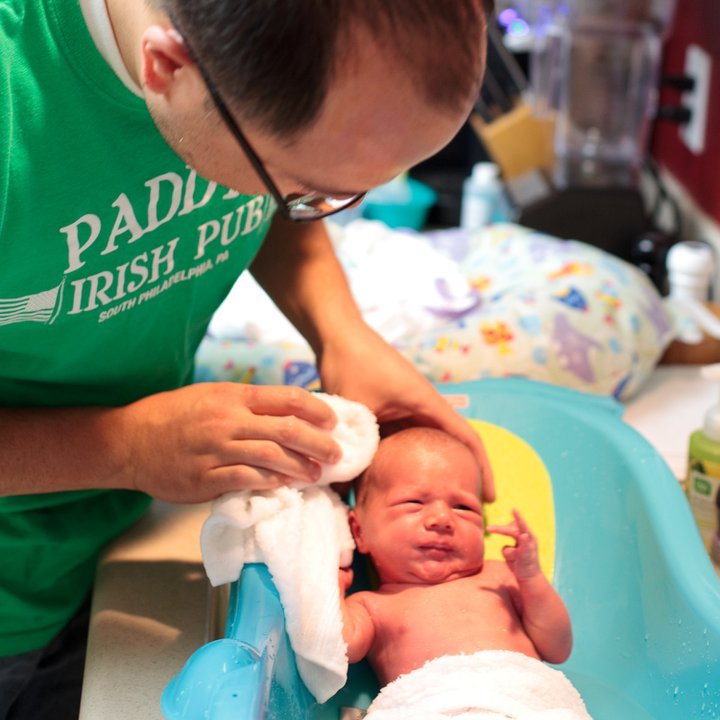 A caregiver in a green shirt gently bathes a newborn baby in a colorful tub, with a soft towel in hand and a warm, cozy home background.