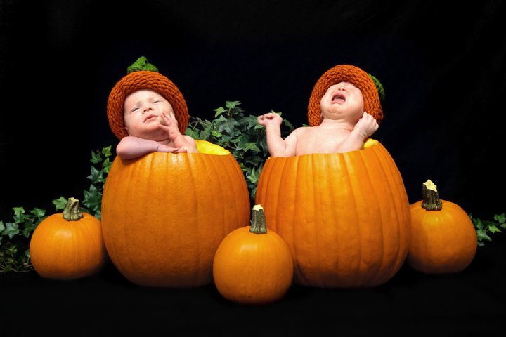 Two babies dressed in pumpkin costumes sitting inside large orange pumpkins with a smaller pumpkin in front of them.
