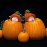 Two sleeping babies wearing orange pumpkin hats nestled inside large pumpkins surrounded by smaller pumpkins.