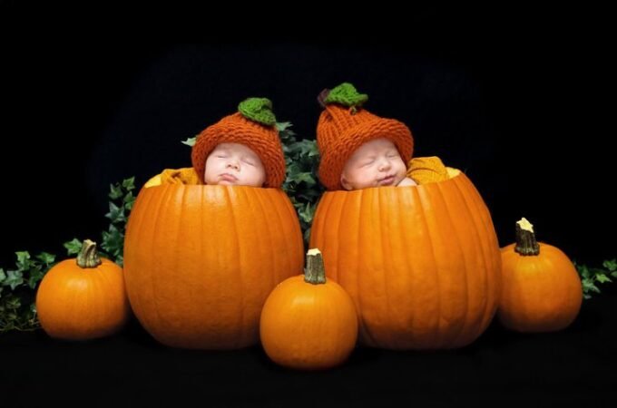 Two sleeping babies wearing orange pumpkin hats nestled inside large pumpkins surrounded by smaller pumpkins.