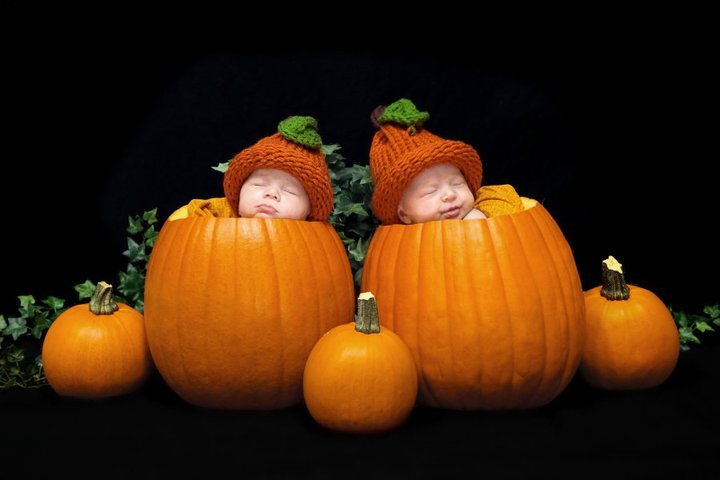 Two sleeping babies wearing orange pumpkin hats nestled inside large pumpkins surrounded by smaller pumpkins.