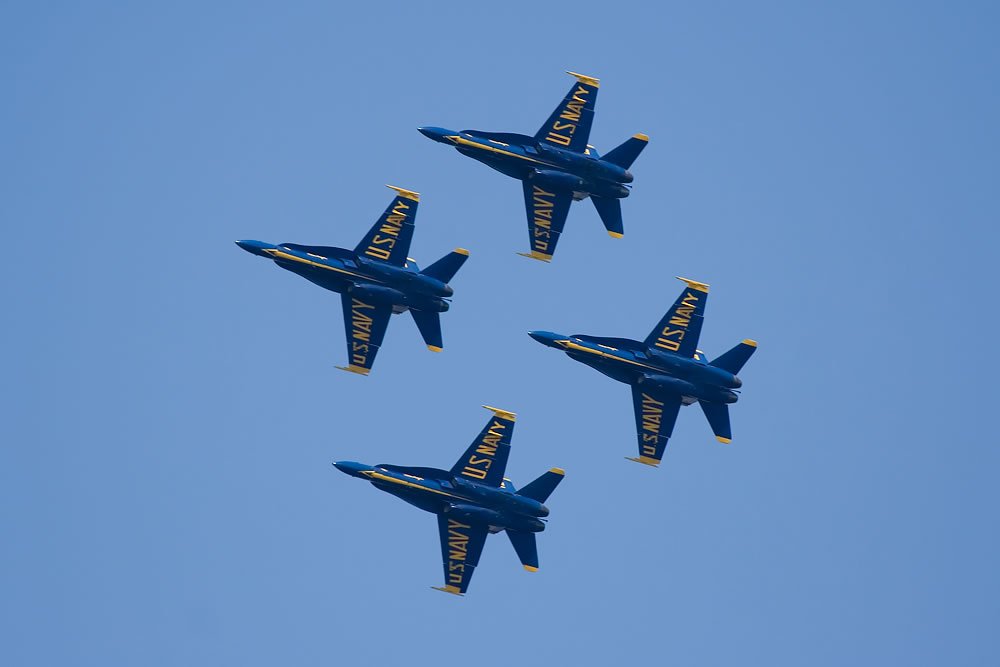 Four Blue Angel jets perform aerial maneuvers with another jet entering the frame from below.