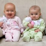 Two babies in pastel rompers sitting on a family sofa, each holding a white stuffed bunny. Smiling babies make for a heartwarming photo moment.