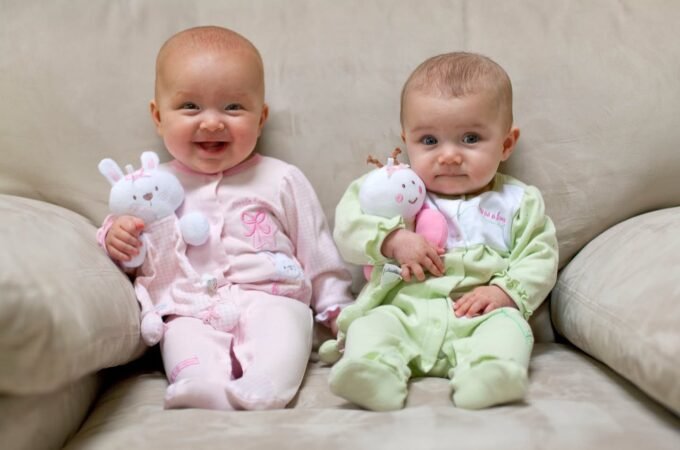 Two babies in pastel rompers sitting on a family sofa, each holding a white stuffed bunny. Smiling babies make for a heartwarming photo moment.