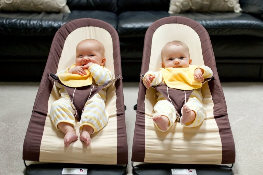 Two babies in matching yellow outfits resting in brown and cream infant bouncers in a living room.