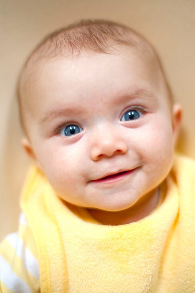 Close up of a smiling baby with bright blue eyes wearing a soft yellow sweater against a warm beige background.