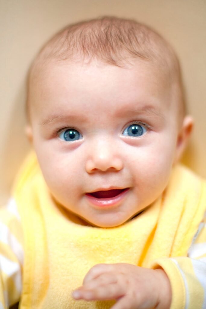 A smiling baby with bright blue eyes wears a cheerful yellow shirt. The close-up captures the joy of early childhood.