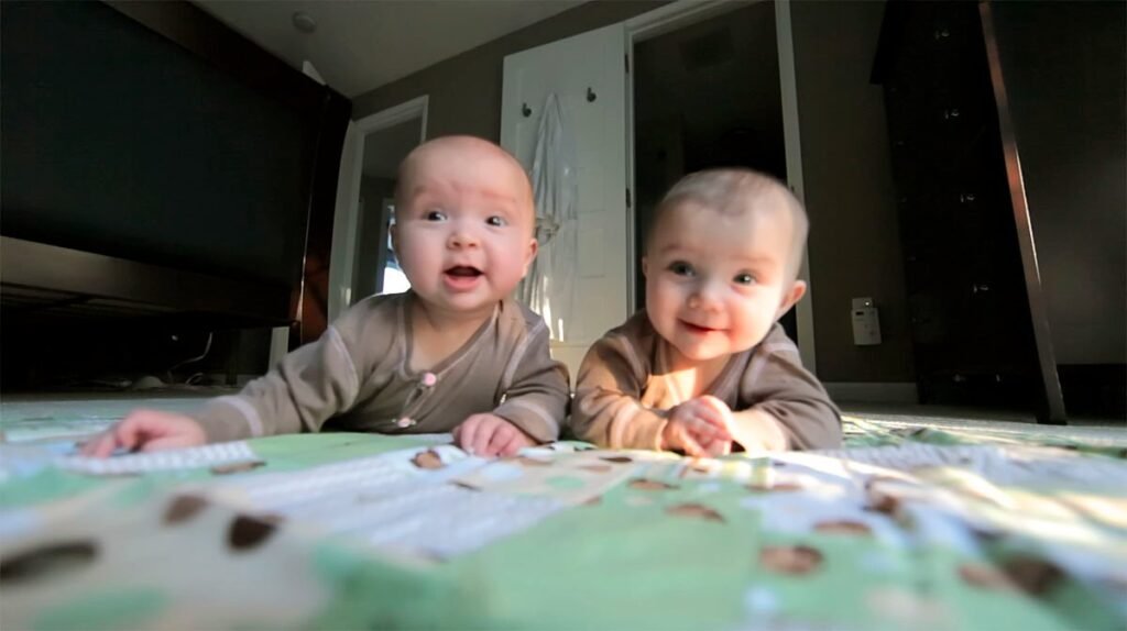 Two babies playing on a patterned blanket in a cozy living room with a TV nearby.