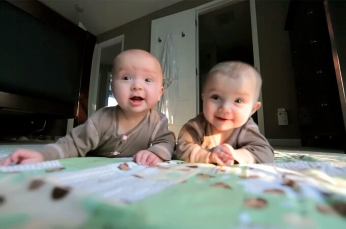 Two babies playing on a patterned blanket in a cozy living room with a TV nearby.