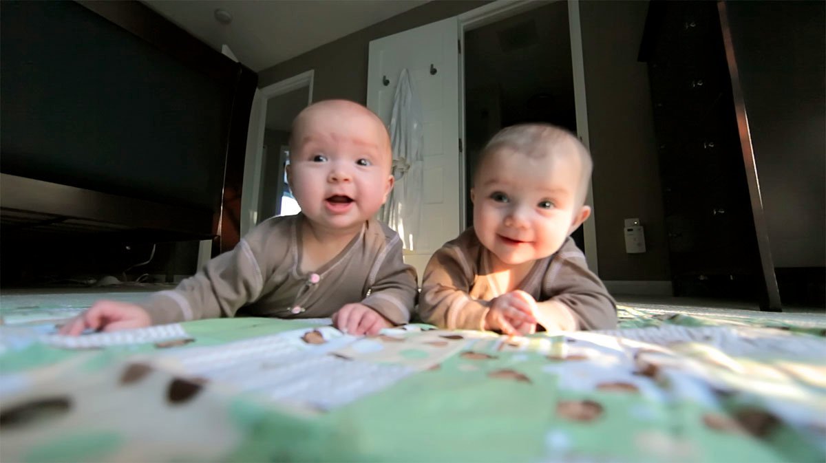 5 months Two babies playing on a patterned blanket in a cozy living room with a TV nearby.
