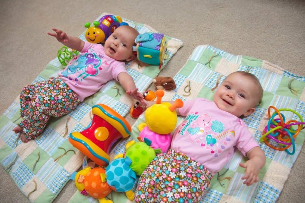 Two babies in matching outfits lie on a soft play mat surrounded by various colorful toys, capturing a moment of pure joy.