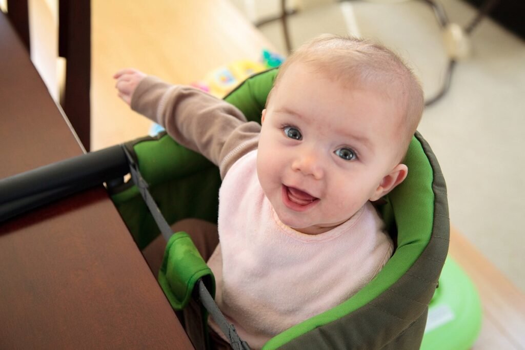 A happy baby with light hair sits in a green high chair, smiling directly at the camera. The background features soft focus indoors.