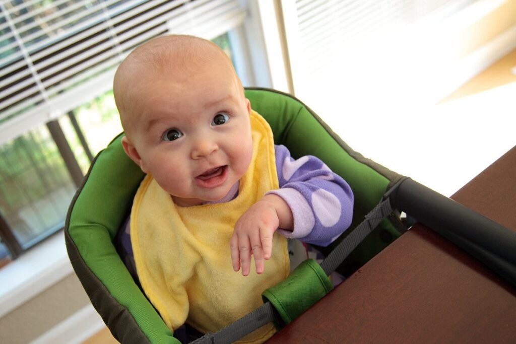 A smiling infant wearing a yellow bib and purple clothes sits in a green high chair. Natural light streams in from a nearby window with blinds.