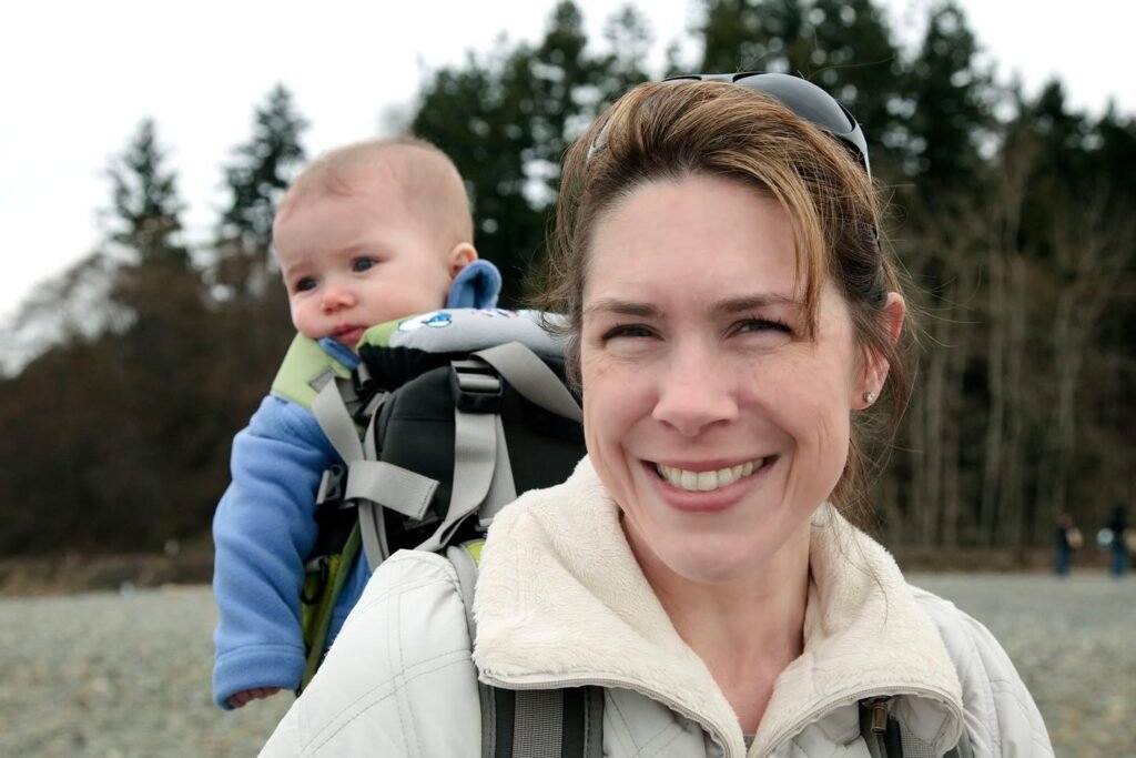 A woman wearing a baby in a back carrier smiles for a photo outdoors. Evergreen trees line the background of this nature scene.